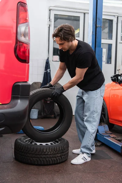 Jeune mécanicien tenant un pneu près des voitures en service — Photo de stock