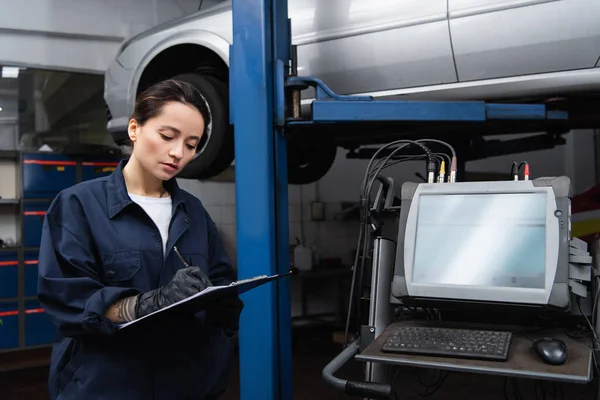 Workwoman writing on clipboard near computer and car in service — Fotografia de Stock