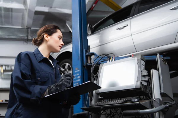 Side view of forewoman writing on clipboard near computer and car in garage — Fotografia de Stock