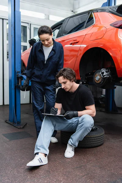 Joven trabajador escribiendo en portapapeles en neumático cerca de colega con destornillador en servicio de coche - foto de stock