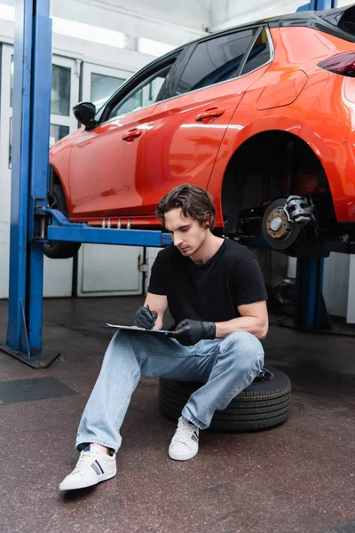 Mechanic in gloves writing on clipboard while sitting on tire near car in garage — Stock Photo