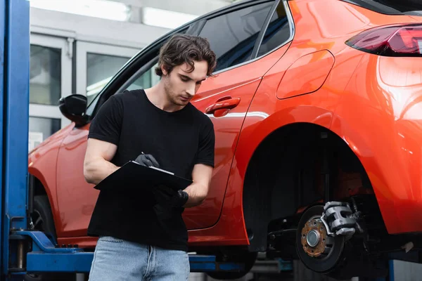 Mecânico em luvas escrevendo na área de transferência perto de automóvel em serviço — Fotografia de Stock