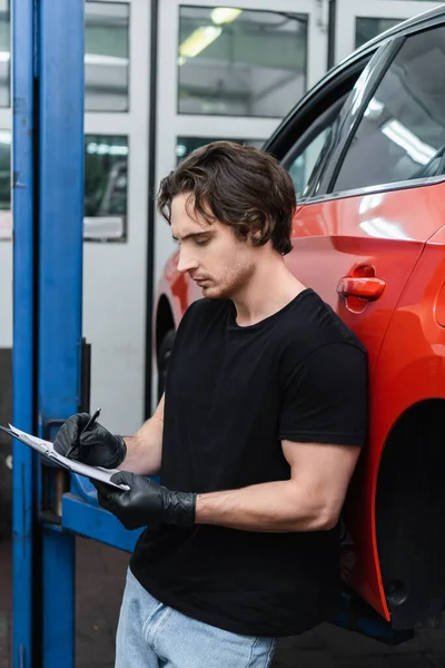 Молодий механік в рукавичках пише на кишені біля машини в гаражі — стокове фото