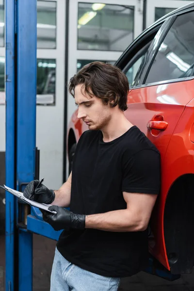 Young mechanic in gloves writing on clipboard near car in garage — Foto stock