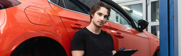 Mechanic holding clipboard and looking at camera near auto in service, banner — Stock Photo
