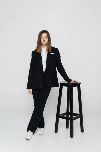 Full length of young stylish woman in suit posing near chair on grey — Fotografia de Stock
