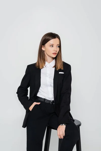 Young stylish woman in suit posing with hand in pocket while sitting chair isolated on grey - foto de stock