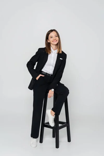 Повна довжина щасливої жінки в костюмі позує рукою в кишені, сидячи на стільці на сірому — стокове фото