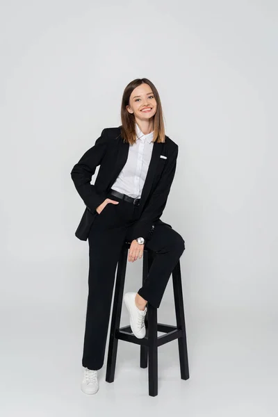 Longitud completa de la mujer feliz en traje posando con la mano en el bolsillo mientras está sentado silla en gris - foto de stock