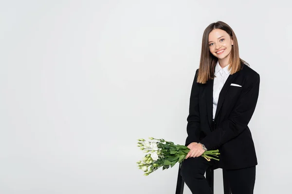 Cheerful woman in suit holding bouquet of flowers and sitting on chair isolated on grey — Foto stock
