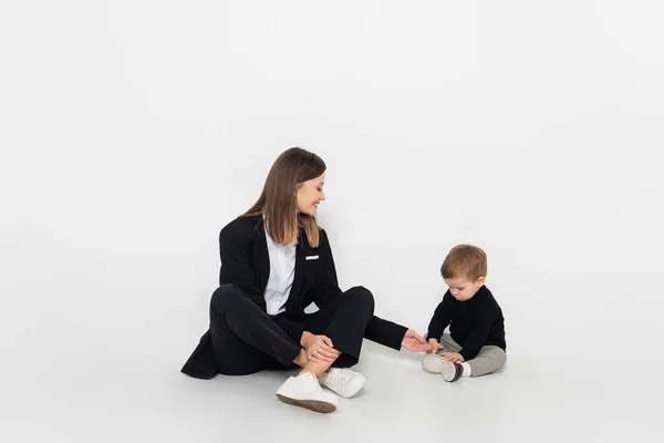 Stylish and happy woman sitting near toddler son on grey - foto de stock