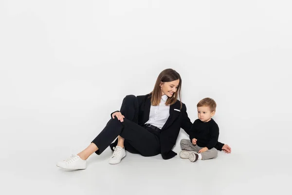 Stylish and cheerful woman sitting near toddler son on grey — стоковое фото