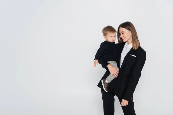 Madre elegante y feliz sosteniendo en brazos hijo pequeño aislado en gris - foto de stock