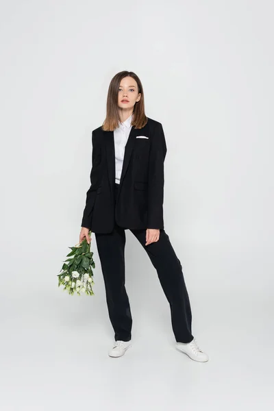 Full length of trendy young woman in black suit holding flowers on grey — стоковое фото