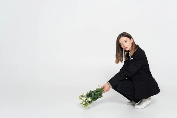 Модна молода жінка в чорному костюмі сидить і тримає білі квіти на сірому — стокове фото