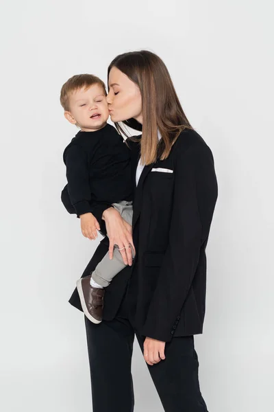 Young woman holding in arms and kissing toddler son isolated on grey — Stockfoto