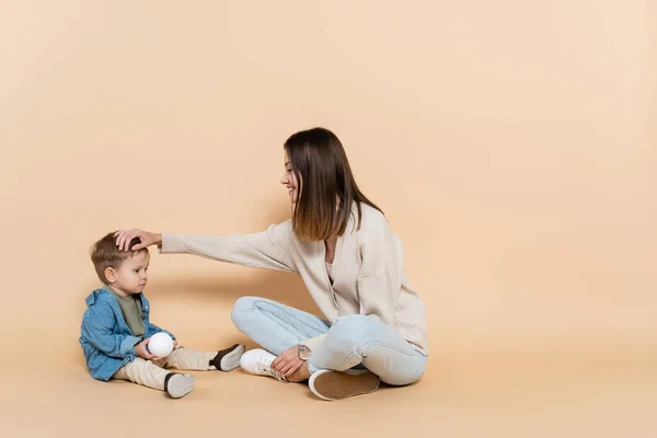 Happy mother sitting and adjusting hair of toddler son on beige — Fotografia de Stock