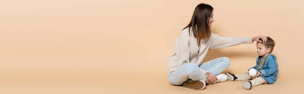 Happy mother sitting and adjusting hair of toddler son on beige, banner — Foto stock