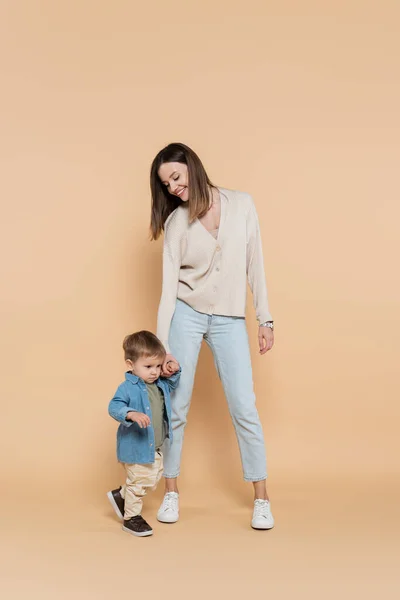 Longitud completa de la madre feliz de pie y tomados de la mano con su hijo pequeño en beige - foto de stock