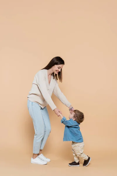 Full length of cheerful mother standing and holding hands with toddler son on beige — Photo de stock