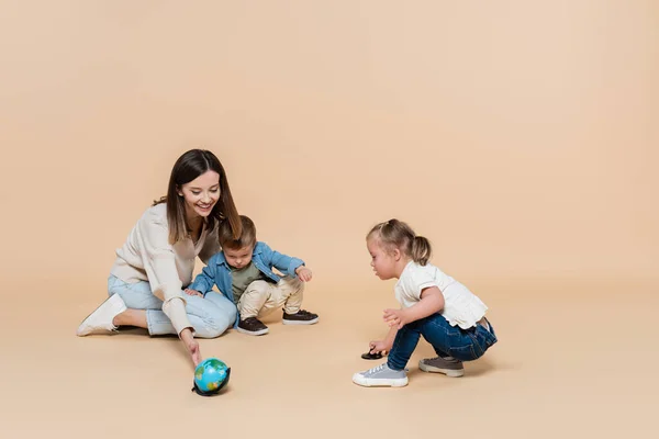 Happy woman playing with globe near toddler boy and girl with down syndrome on beige — Stock Photo