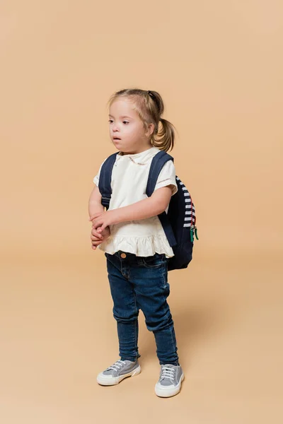 Girl with down syndrome standing with backpack on beige — Stock Photo