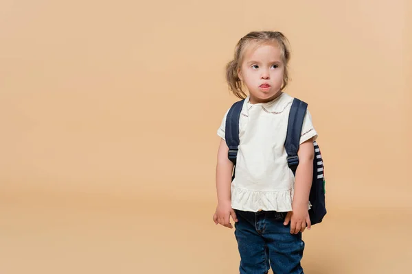 Дитина з синдромом вниз стирчить язиком, стоячи з рюкзаком на бежевому — стокове фото
