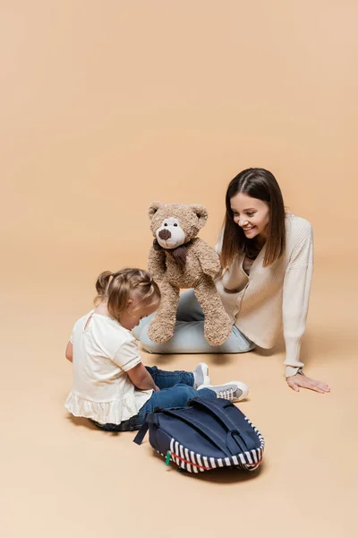 Щаслива мати тримає плюшевого ведмедя біля дівчини з синдромом Дауна і рюкзаком на бежевому — стокове фото
