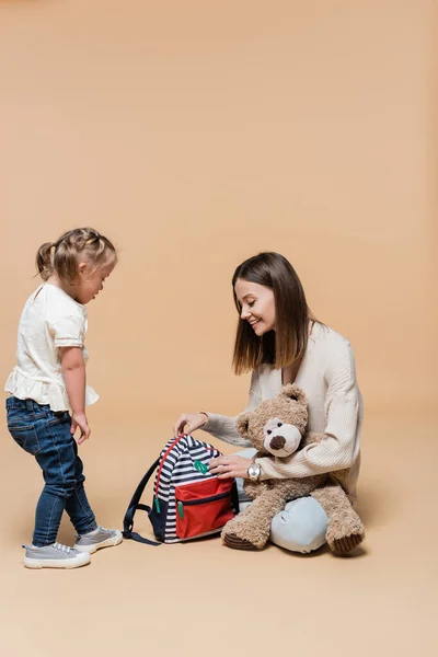 Happy mother holding teddy bear and backpack near girl with down syndrome on beige — Stockfoto