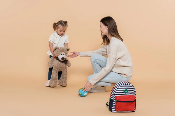 Happy mother holding small globe near girl with down syndrome hugging teddy bear on beige — Stock Photo