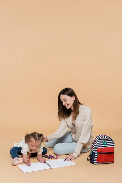 Cheerful mother sitting near girl with down syndrome drawing near colorful pencils and backpack on beige — Stockfoto