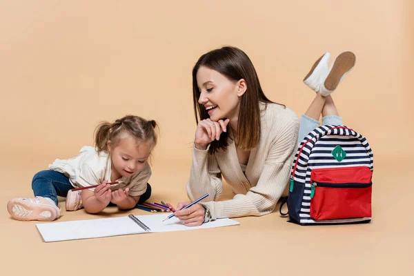 Positive mother lying near child with down syndrome drawing near colorful pencils and backpack on beige — Photo de stock