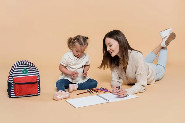 Радісна мати лежить біля дитини з синдромом вниз малює біля барвистих олівців і рюкзака на бежевому — стокове фото