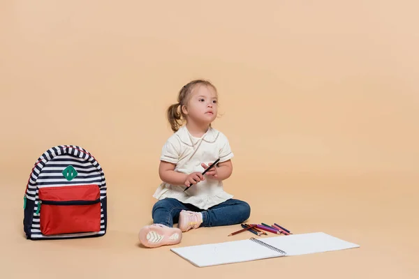 Kid with down syndrome sitting near colorful pencils and backpack on beige — Stockfoto