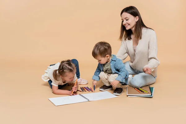 Happy woman sitting near toddler boy and girl with down syndrome drawing on beige — стоковое фото
