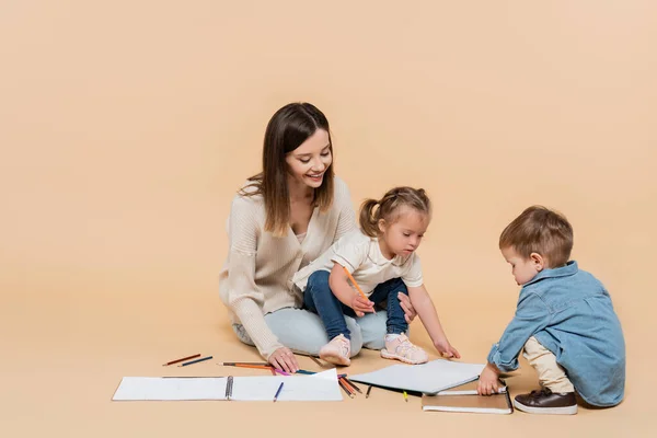 Щаслива мати сидить біля дівчини з синдромом Дауна, хлопчик-малюк і барвисті олівці на бежевому — стокове фото