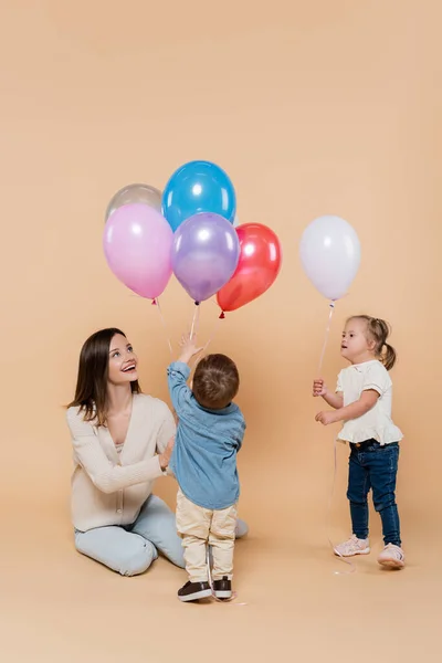 Happy mother sitting near girl with down syndrome and toddler boy holding colorful balloons on beige — Foto stock