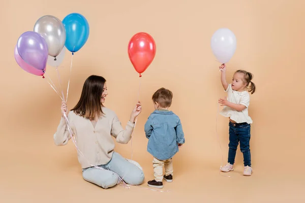 Cheerful mother sitting near girl with down syndrome and toddler boy holding colorful balloons on beige — Photo de stock