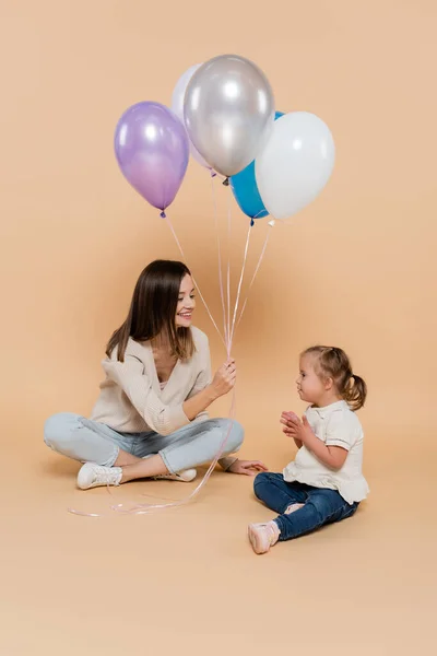 Positive woman sitting near girl with down syndrome while holding colorful balloons on beige — Photo de stock