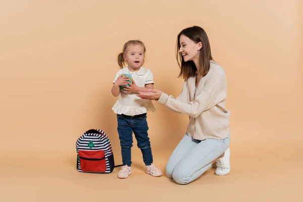 Chica con síndrome de Down sosteniendo pequeño globo terráqueo cerca feliz madre y mochila en beige - foto de stock