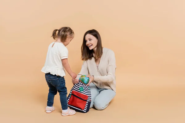 Mujer feliz sosteniendo pequeño globo y mochila mientras mira a niño con síndrome de Down en beige - foto de stock