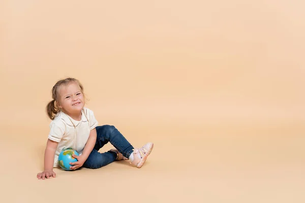 Щаслива дитина з синдромом Дауна сидить з невеликою глобусом на бежевому — стокове фото
