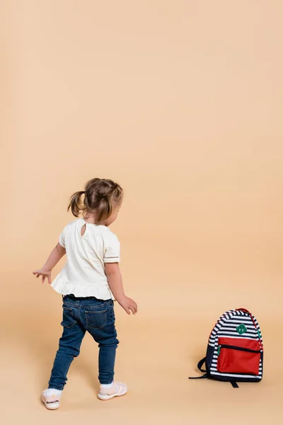 Kid with down syndrome walking near backpack on beige — стоковое фото