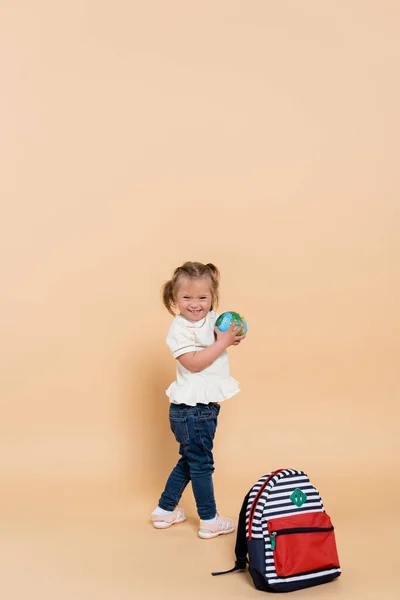 Щаслива дитина з синдромом Дауна, що тримає невеликий глобус біля рюкзака на бежевому — стокове фото