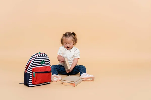 Kid with down syndrome sitting near books and backpack on beige — Photo de stock