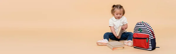 Kid with down syndrome sitting near books and backpack on beige, banner — стоковое фото