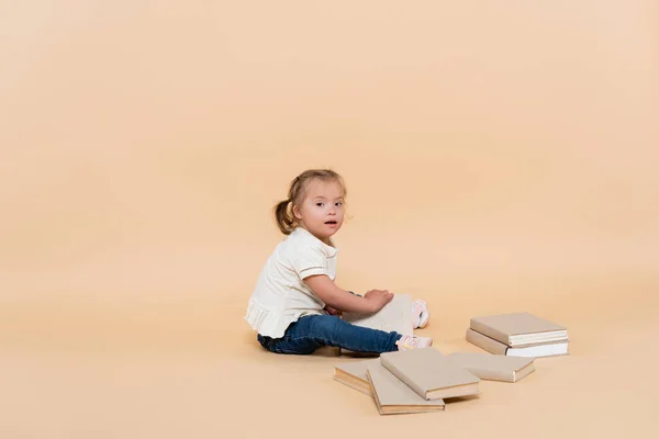 Chica con síndrome de Down sentado cerca de libros en beige - foto de stock