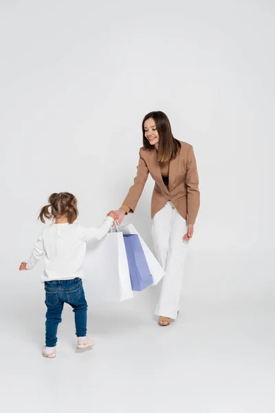 Happy and stylish mother holding shopping bags while looking at kid with down syndrome on grey — стоковое фото