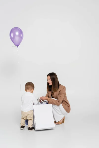Feliz madre sosteniendo bolsas de compras cerca de niño pequeño hijo y globo en gris - foto de stock