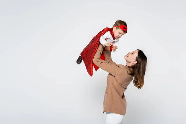Feliz madre levantando hijo pequeño en traje de superhéroe aislado en gris - foto de stock